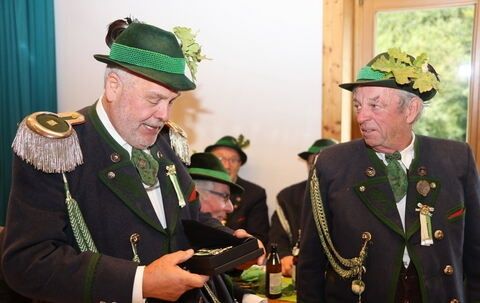 Eine Gruppe von Männern in traditionellen grünen Hüten und Uniformen unterhalten sich, einer hält eine Schachtel und der andere lächelt. Sie befinden sich in einem Raum mit Holzwänden und Blick auf Bäume im Freien.