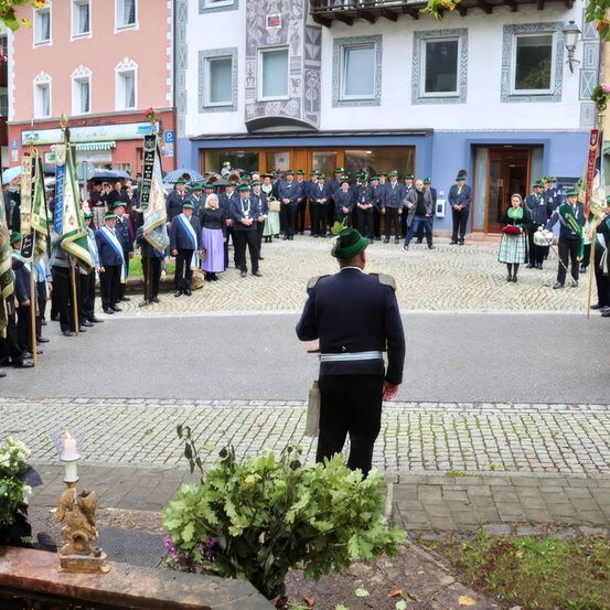 Eine Gruppe von Menschen in Uniformen steht vor einem Gebäude mit vielen Fenstern. Einige tragen Fahnen. Eine Person mit einem grünen Hut steht im Vordergrund.