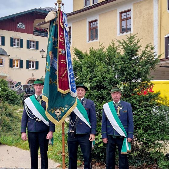 Drei Männer in traditioneller bayerischer Uniform stehen vor einem Fahnenmast. Hinter ihnen befinden sich Büsche und ein gelbes Gebäude mit mehreren Fenstern.