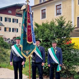 Drei Männer in traditioneller bayerischer Uniform stehen vor einem Fahnenmast. Hinter ihnen befinden sich Büsche und ein gelbes Gebäude mit mehreren Fenstern.