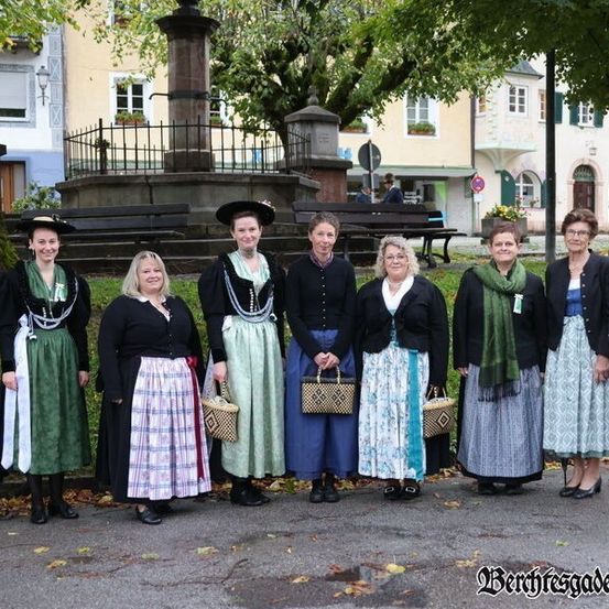 Eine Gruppe von Menschen, gekleidet in traditioneller Kleidung, steht vor einem Brunnen, lächelt und posiert für ein Foto. Die Umgebung umfasst Gebäude, Bäume und eine Bank.