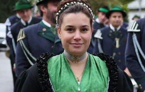 Eine Frau in einem traditionellen grünen und schwarzen Kleid mit Perlenkette steht vor einer Gruppe von Männern in gleichen Uniformen.