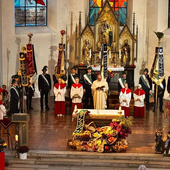 Eine religiöse Zeremonie findet in einer Kirche statt, an der eine Gruppe von Menschen teilnimmt. Ein Altar, Kerzen und Blumen sind vorhanden. Die Menschen tragen traditionelle Kleidung.
