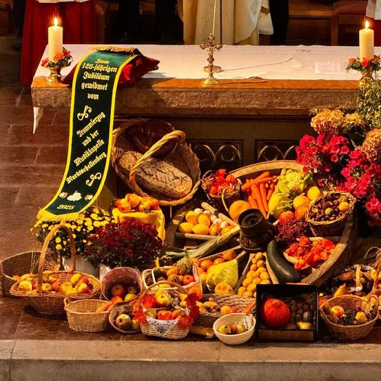 Ein Tisch mit verschiedenen Früchten und Gemüse ist aufgestellt, umgeben von Körben. Eine grüne Fahne trägt den Text '125 Jahre Jubiläum'. Dahinter bedeckt ein weißes Tuch den Tisch. Zwei Kerzen sind angezündet.