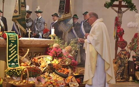 Ein Priester betet vor einem mit Früchten und Blumen gefüllten Altar, umgeben von uniformierten Männern und einem Kreuz.