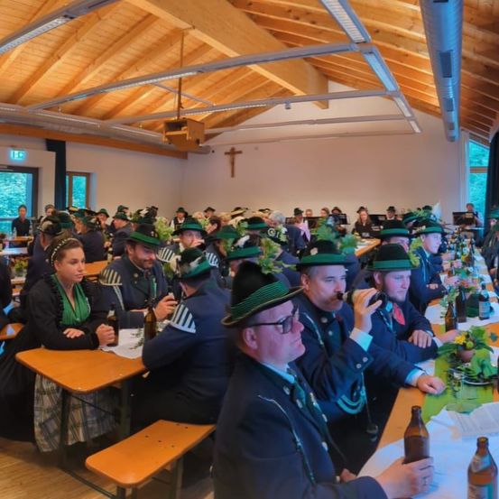 In einem hölzernen Raum sind Menschen versammelt, die grüne Hüte tragen. Einige trinken aus Flaschen, und es gibt Tische mit Pflanzen und Flaschen. Der Raum hat Holzdecken und weiße Wände.