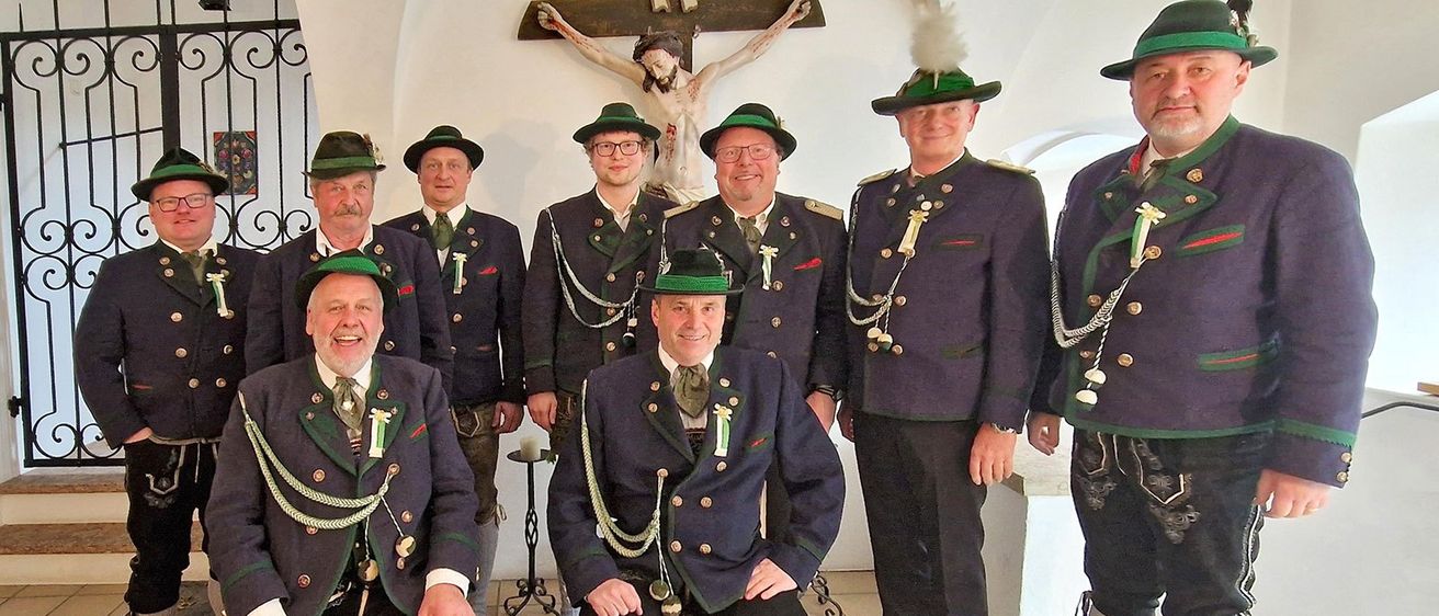 Eine Gruppe von Männern in traditioneller bayerischer Kleidung posiert für ein Foto vor einem Kruzifix. Sie alle lächeln und einige sitzen.