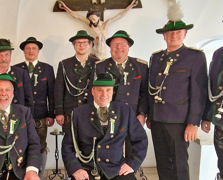 Eine Gruppe von Männern in traditioneller bayerischer Kleidung posiert für ein Foto vor einem Kruzifix. Sie alle lächeln und einige sitzen.