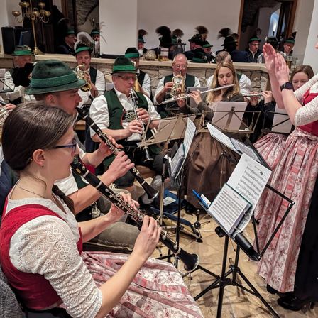 Eine Gruppe von Musikern tritt in einem Raum auf und trägt traditionelle Kleidung. Zu den Musikern gehören eine Frau, die Flöte spielt, ein Mann, der Klarinette spielt, und andere, die Trompeten und Posaunen spielen. Vor ihnen stehen Notenständer mit Notenblättern.