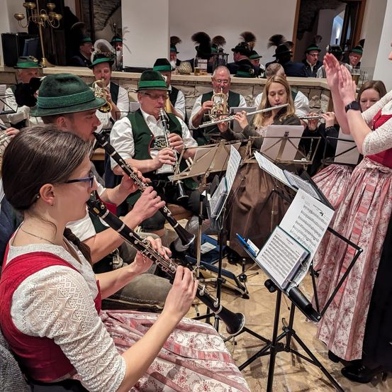 Eine Gruppe von Musikern tritt in einem Raum auf und trägt traditionelle Kleidung. Zu den Musikern gehören eine Frau, die Flöte spielt, ein Mann, der Klarinette spielt, und andere, die Trompeten und Posaunen spielen. Vor ihnen stehen Notenständer mit Notenblättern.