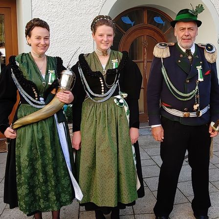 Drei Personen in traditioneller Kleidung stehen vor einem Gebäude mit einer Holztür und Glasfenstern. Die Frau auf der linken Seite hält ein Horn. Die andere Frau und der Mann tragen Uniformen mit Verzierungen.