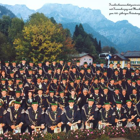 Eine große Gruppe von Musikern in Uniform posiert für ein Foto mit Bergen im Hintergrund. Sie halten verschiedene Instrumente und sind in Reihen angeordnet.