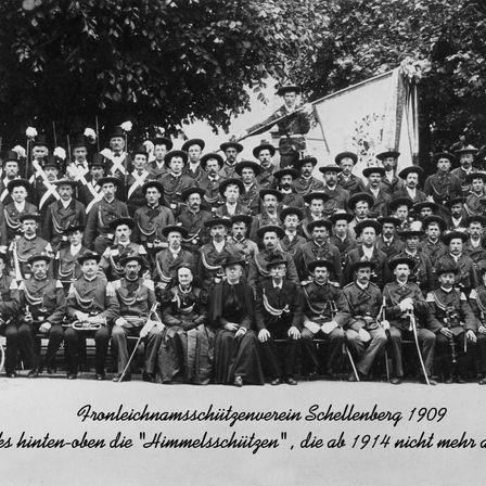 Schwarz-Weiß-Foto einer Gruppe von uniformierten Soldaten und Soldatinnen mit Hüten, die 1909 in Schellenberg stehen und auf einer Bank sitzen.