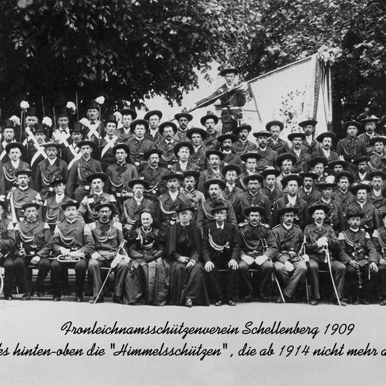Schwarz-Weiß-Foto einer Gruppe von uniformierten Soldaten und Soldatinnen mit Hüten, die 1909 in Schellenberg stehen und auf einer Bank sitzen.