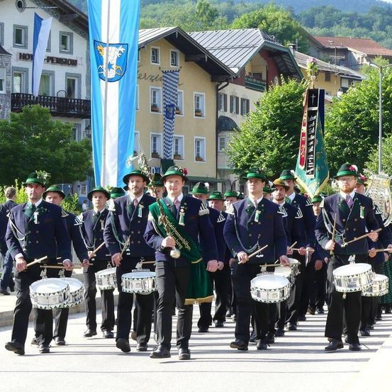 Eine Marschkapelle in traditionellen Uniformen und grünen Hüten spielt Trommeln und folgt Fahnen auf einer Straße. Im Hintergrund befinden sich Gebäude und Bäume.