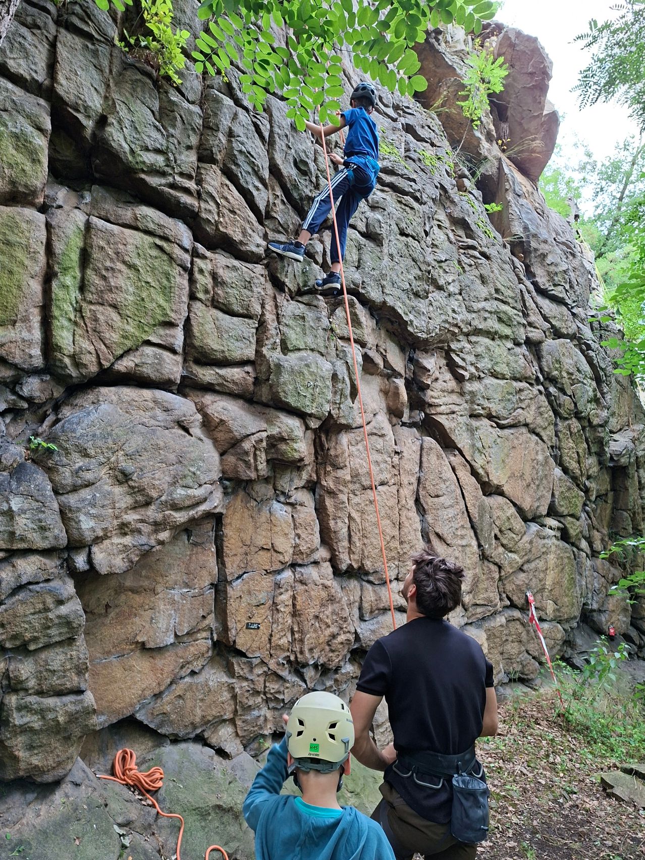 Eine Person klettert an einem Felsen, gesichert durch ein Seil, während eine andere Person in der Nähe zuschaut. Der Kletterer trägt einen Helm, und die Felswand ist rau.