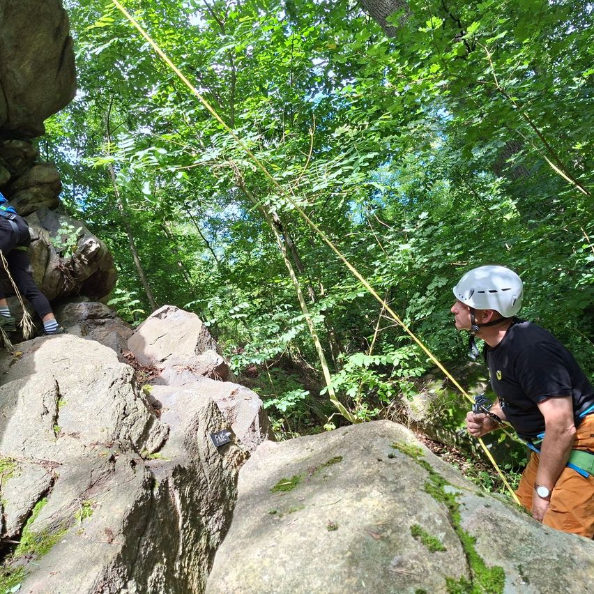 Zwei Personen klettern in einem Wald auf Felsen. Einer trägt einen weißen Helm, während der andere ein Seil benutzt. Beide sind mit Rucksäcken ausgestattet.