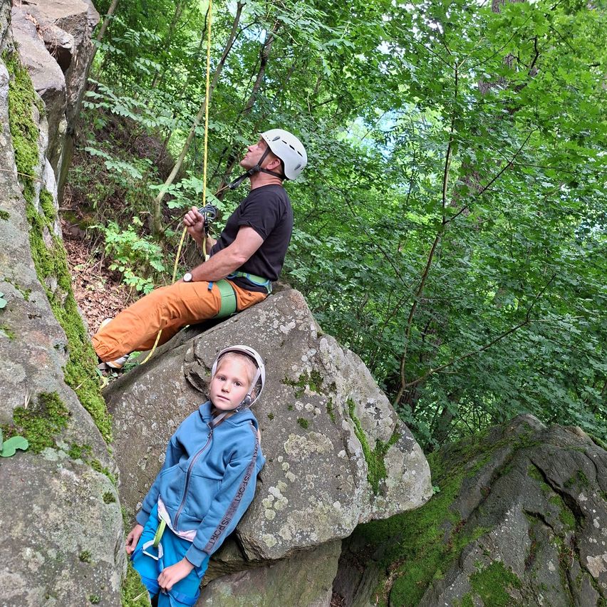 Ein Mann und ein Kind klettern in einem Wald. Der Mann sitzt auf einem großen Felsen und bereitet sich auf das Klettern vor, während das Kind auf dem Boden ruht.