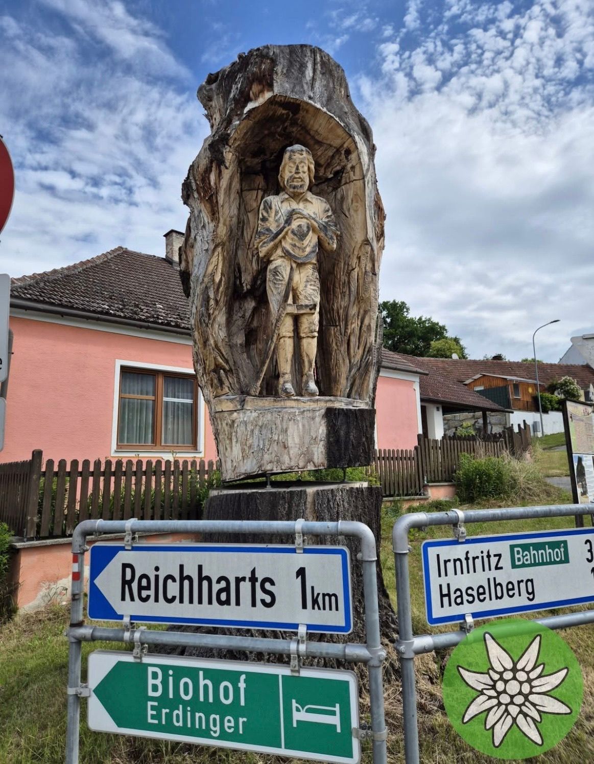 Eine Holzstatue eines Mannes ist in einen Baumstumpf geschnitzt. Die Statue wird von Schildern umgeben, die Reicharts 1km und Irnfritz Bahnhof 3km anzeigen. Im Hintergrund steht ein rosafarbenes Haus mit Fenster und Zaun.