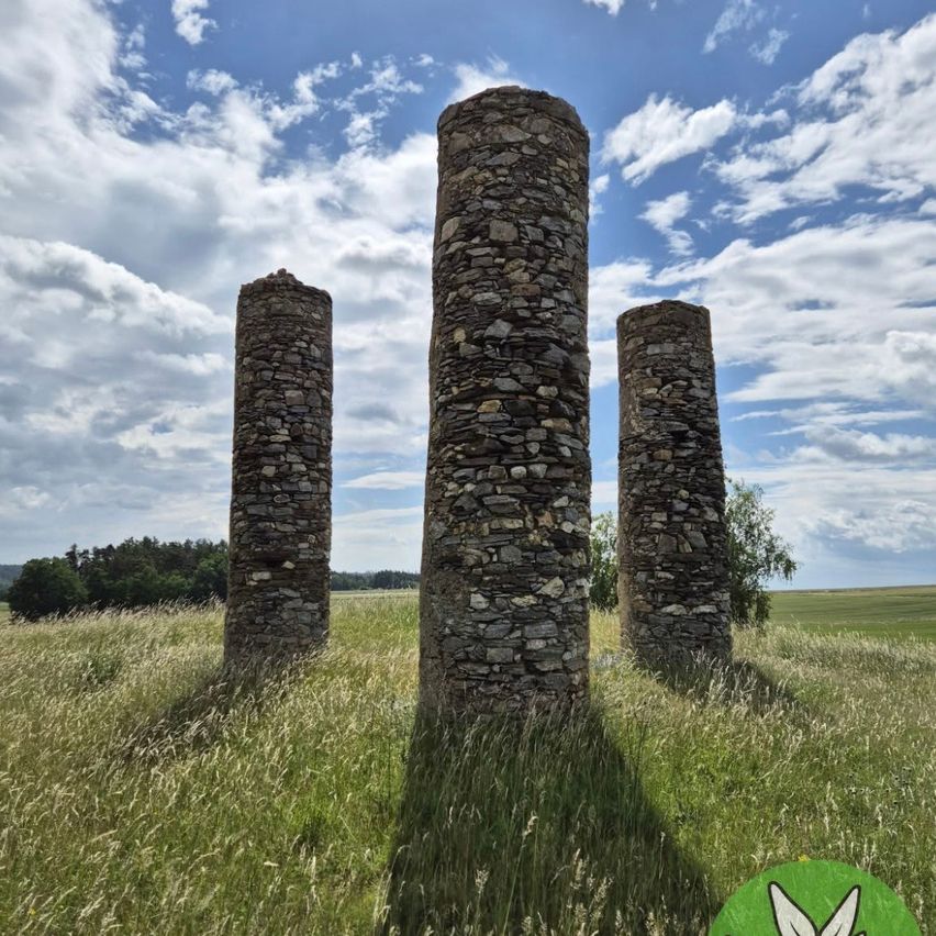 Drei Steinsäulen in einem Grasfeld unter einem blauen Himmel mit Wolken.