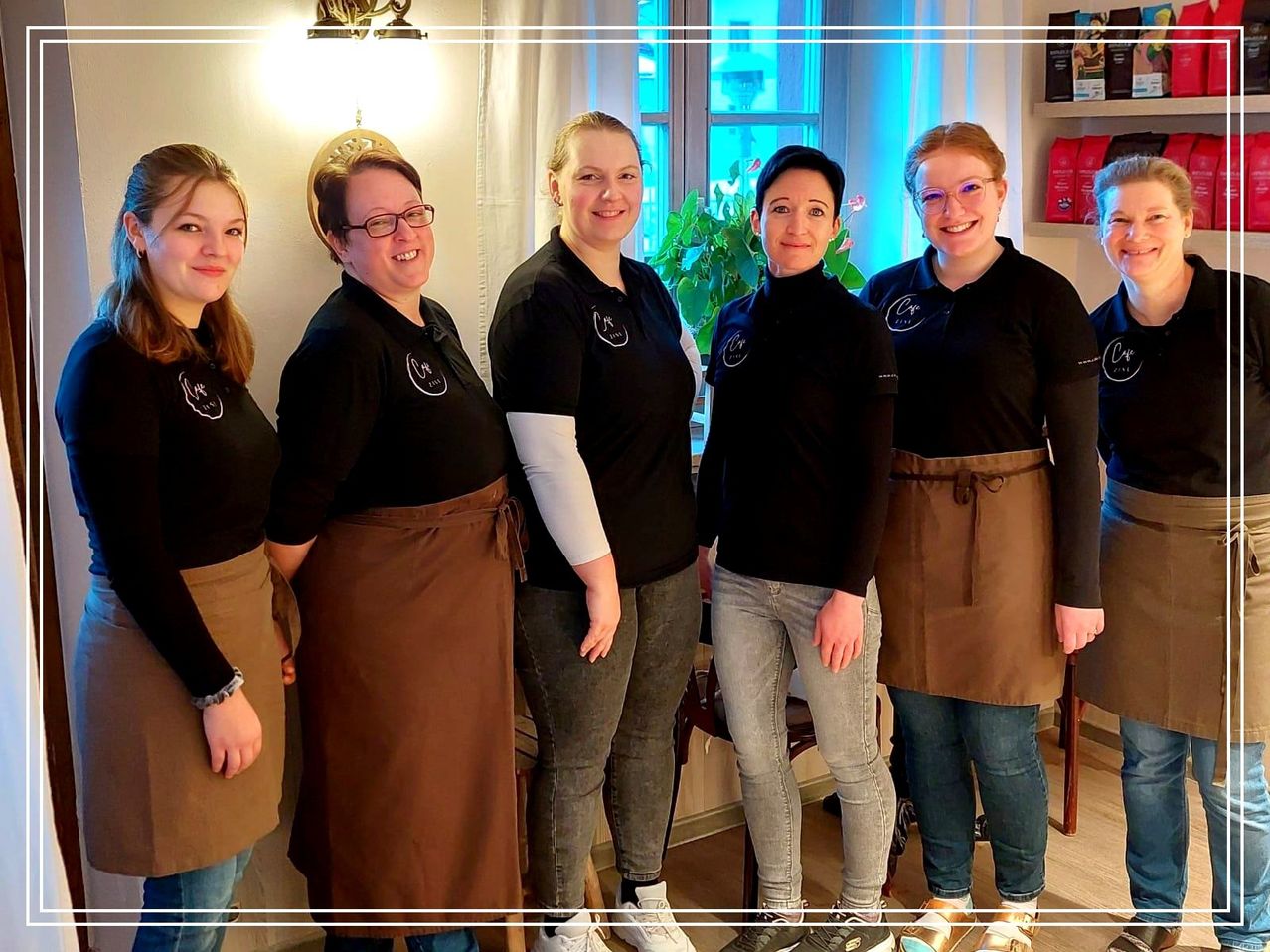 Fünf Frauen stehen gemeinsam in einem Raum. Sie tragen passende schwarze Shirts und braune Schürzen. Sie alle lächeln und scheinen für ein Foto zu posieren. Hinter ihnen befindet sich ein Regal mit verschiedenen Gegenständen.