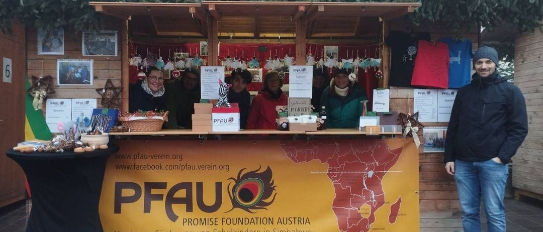 Eine Gruppe von Menschen steht hinter einem hölzernen Tresen mit einem Banner, auf dem 'PFAU Promise Foundation Austria' steht. Das Banner zeigt eine Karte von Afrika. Der Tresen ist mit Gegenständen gefüllt und es herrscht eine festliche Atmosphäre.