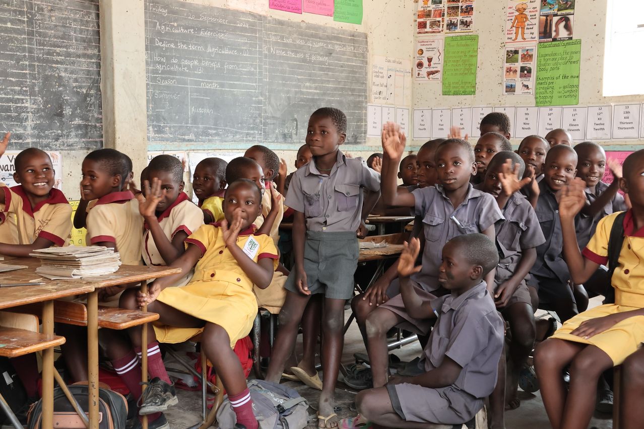 Ein Klassenzimmer voller Kinder in Uniformen, die ihre Hände heben. Ein Junge in einem gelben Kleid steht auf. Ein Tafel mit Notizen über Landwirtschaft und Jahreszeiten steht hinter ihnen.