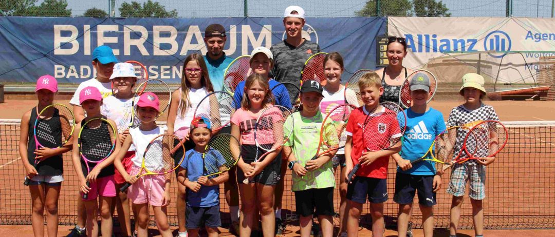 Eine Gruppe von Kindern und Erwachsenen posiert für ein Foto auf einem Tennisplatz mit Schlägern. Dahinter ist ein Zaun mit einem Banner zu sehen, auf dem 'Baum' steht.