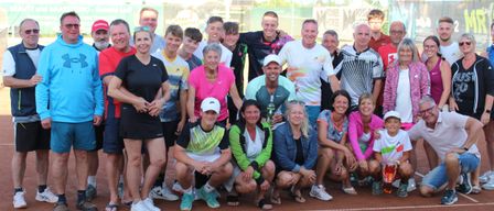 Eine Gruppe von Menschen posiert für ein Foto auf einem Tennisplatz. Sie sind in verschiedenen Farben gekleidet und einige tragen Hüte. Der Platz hat weiße Linien und im Hintergrund befinden sich Zäune und Banner.