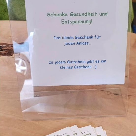 Ein durchsichtiger Kunststoffständer hält eine weiße Karte mit blauem Text, die ein Geschenk für jeden Anlass verspricht und Gesundheit und Entspannung bietet. Unter der Karte sind mehrere kleine Karten zu sehen.