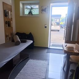 Ein Raum mit einem Massagetisch, einem Topfpflanzenfenster und einer offenen Tür, die zu einem Parkplatz führt.