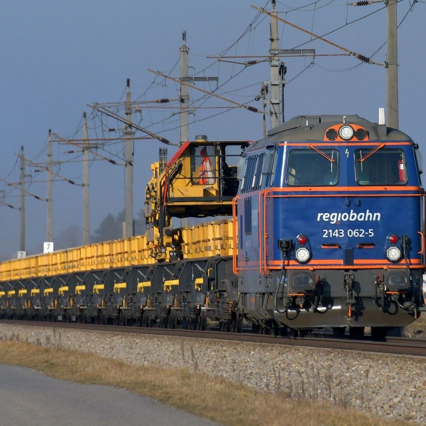 Ein blauer und orangefarbener Zug fährt auf den Gleisen und zieht einen großen gelben und schwarzen Frachtwagen. Oberleitungsmasten verlaufen entlang der Seite der Gleise.