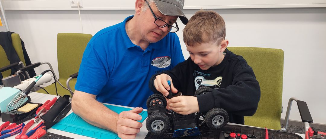 Ein älterer Mann hilft einem jungen Jungen, die Schrauben an einem Spielzeugauto zu justieren. Der Junge trägt ein schwarzes Shirt und der Mann ein blaues Polohemd mit Logo.