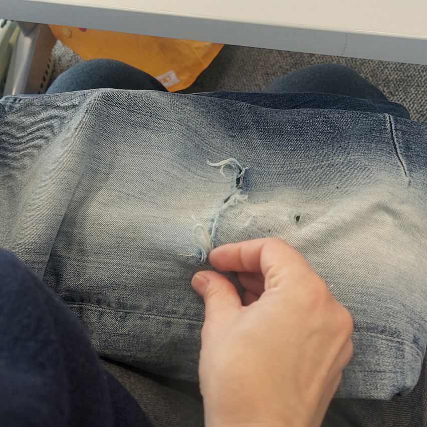 Eine Person repariert eine Jeanstasche mit einer Nadel. Die Jeans sind hellblau und verwaschen. Die Hand der Person hält die Jeans am Rand.