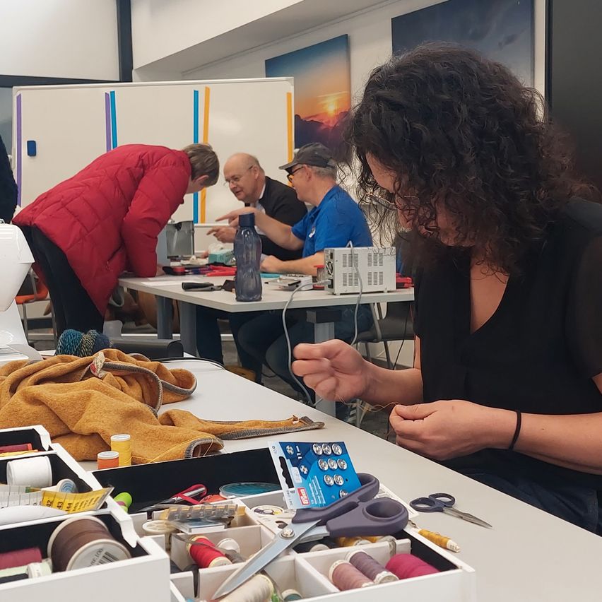 Eine Gruppe von Menschen befindet sich in einem Workshop, näht und arbeitet an einem Projekt. Ein Tisch ist mit verschiedenen Nähzubehör gefüllt.