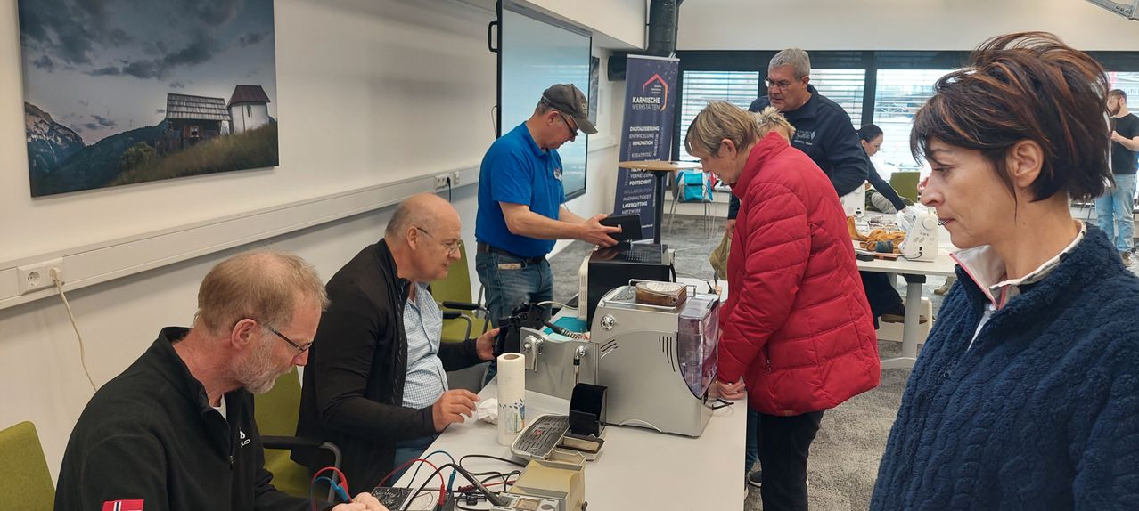 Zwei Männer untersuchen ein Gerät an einem Tisch. Dahinter bedient ein Mann eine Maschine. Im Hintergrund spricht ein Mann mit einer Frau. An der Wand befindet sich eine Tafel und ein Banner.