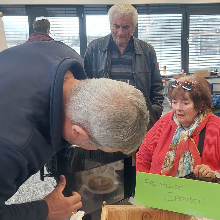 Ein älterer Mann betrachtet einen Kuchen in einer Glasvitrine, während ein anderer Mann und eine Frau in einer roten Jacke in der Nähe stehen. Das Schild sagt 'Freiwillige Spenden'.