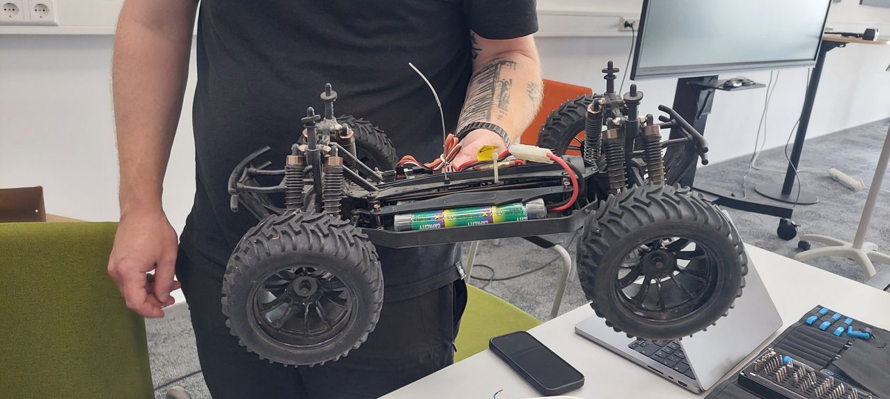 Eine Person hält ein Spielzeugauto mit sichtbarer Batterie. Das Auto hat vier Reifen und eine Fernbedienung liegt auf dem Tisch.