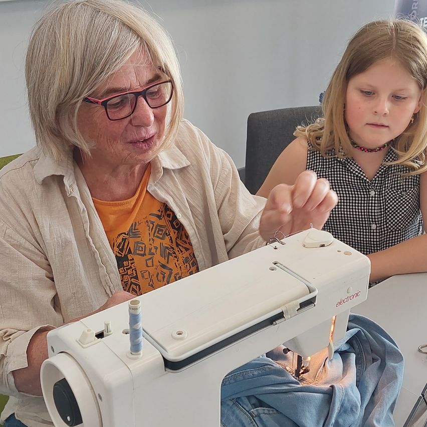 Eine ältere Frau mit Brille benutzt eine Nähmaschine, während ein junges Mädchen neben ihr sitzt. Sie befinden sich beide in einem Raum.
