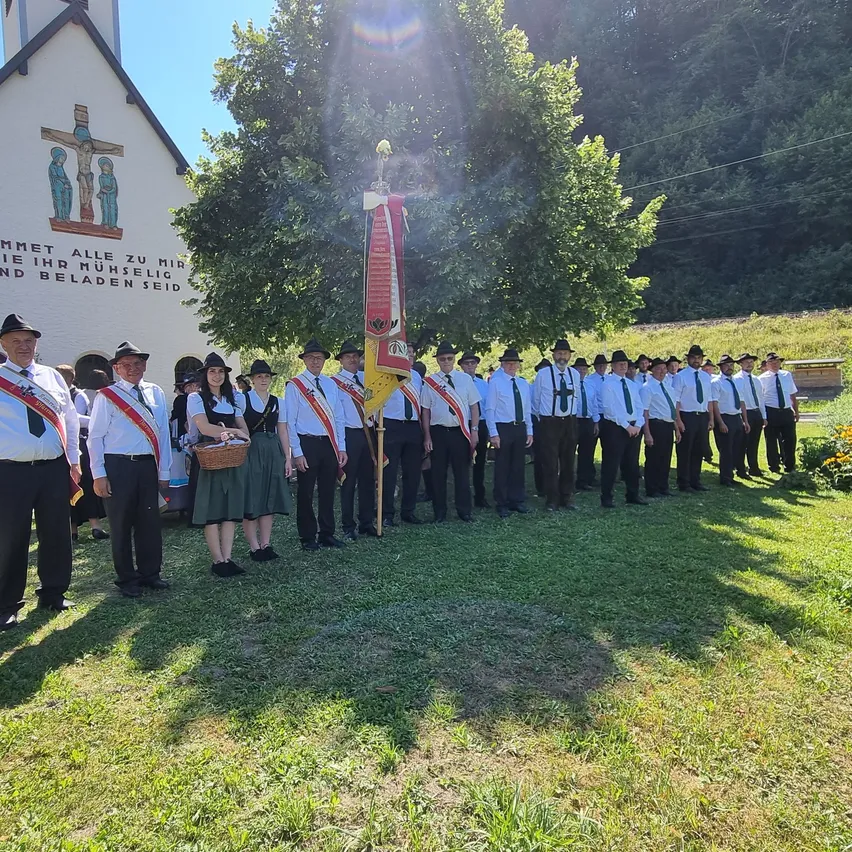 Eine Gruppe von Menschen in traditioneller Kleidung steht vor einer Kirche, hält eine Fahne und einen Korb.