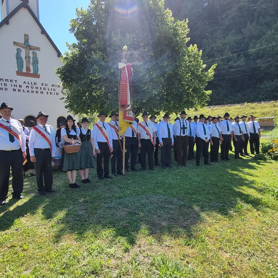 Eine Gruppe von Menschen in traditioneller Kleidung steht vor einer Kirche, hält eine Fahne und einen Korb.