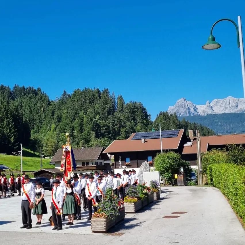 Eine Gruppe von Menschen in traditioneller Kleidung steht in einer Reihe vor einem Gebäude mit Solarmodulen auf dem Dach. Im Hintergrund befindet sich eine Bergkette und ein klarer blauer Himmel.