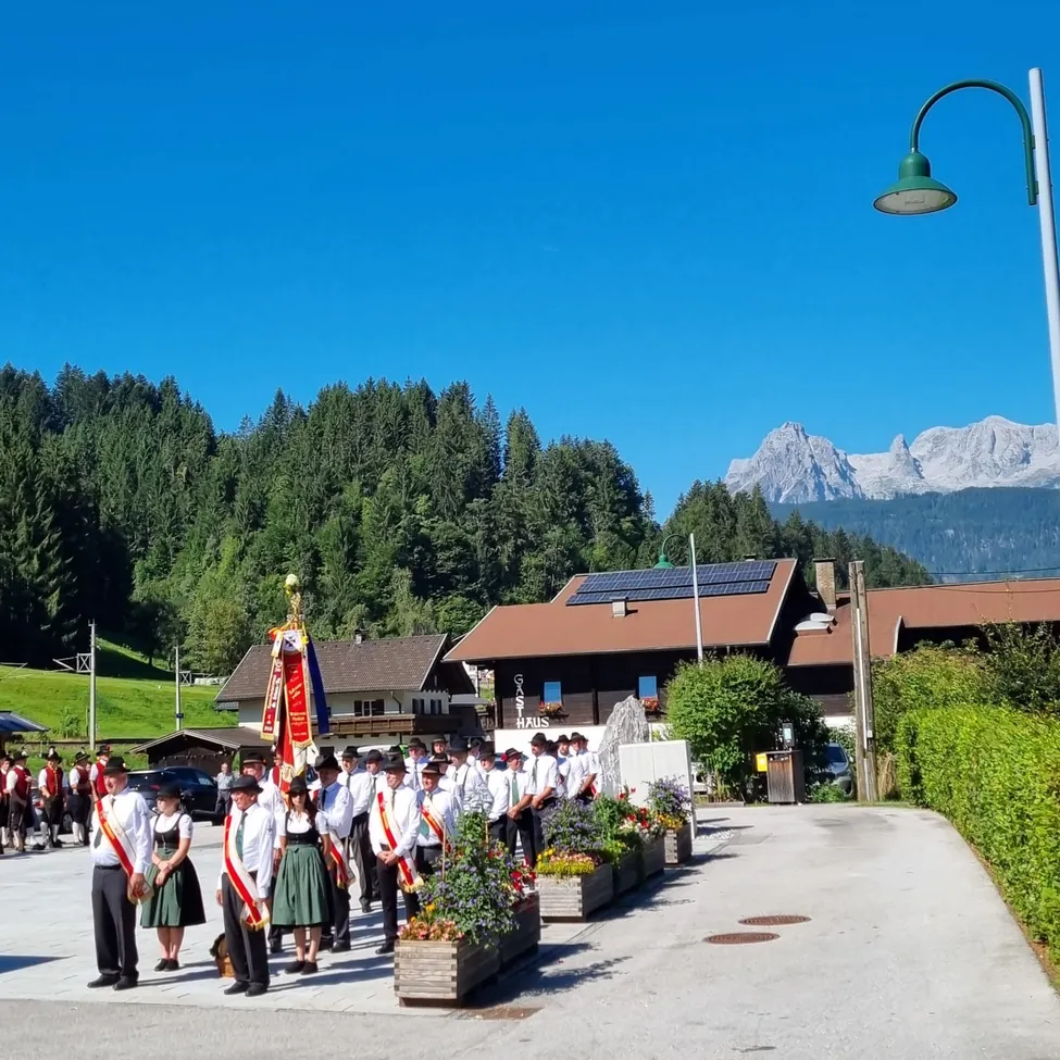 Eine Gruppe von Menschen in traditioneller Kleidung steht in einer Reihe vor einem Gebäude mit Solarmodulen auf dem Dach. Im Hintergrund befindet sich eine Bergkette und ein klarer blauer Himmel.