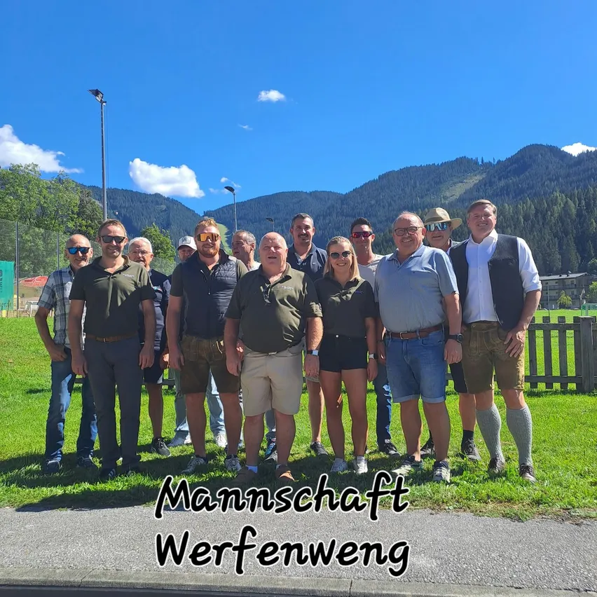 Eine Gruppe von Menschen in lässiger Sommerkleidung, die auf einem Grasfeld mit Bergen im Hintergrund stehen. Unten steht der Text 'Mannschaft Werfenweg'.