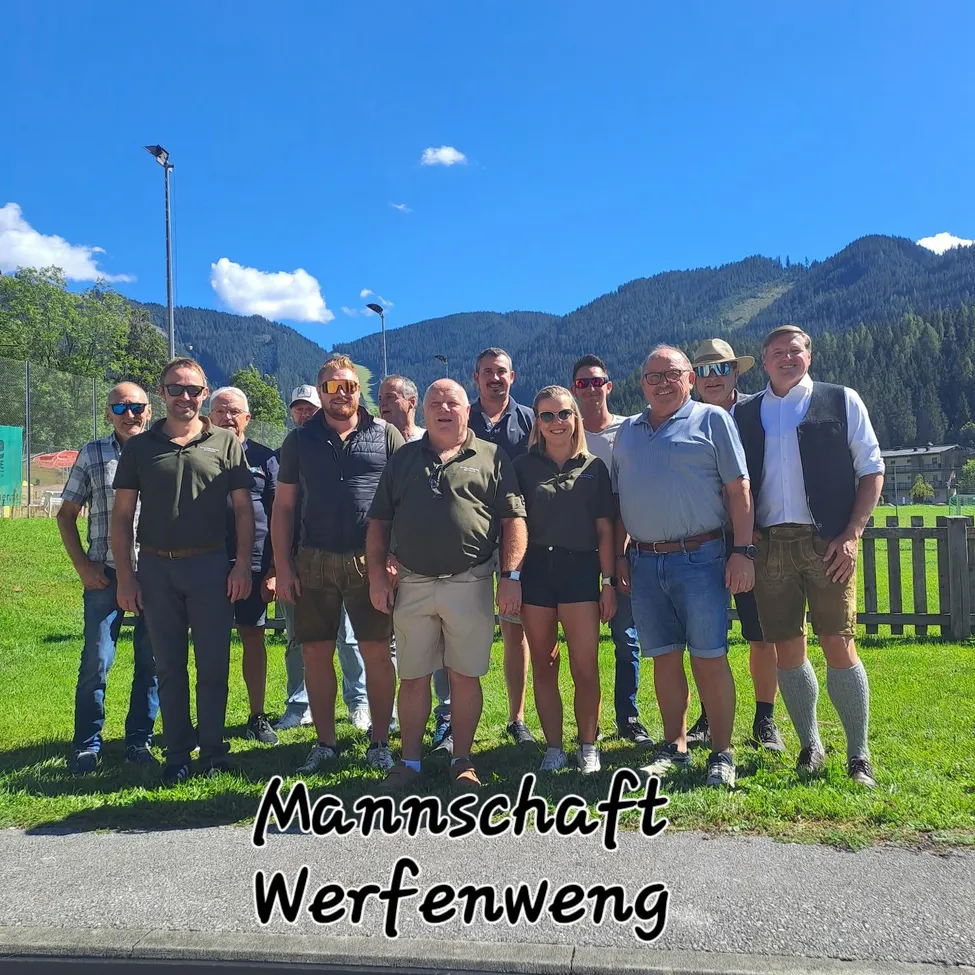 Eine Gruppe von Menschen in lässiger Sommerkleidung, die auf einem Grasfeld mit Bergen im Hintergrund stehen. Unten steht der Text 'Mannschaft Werfenweg'.