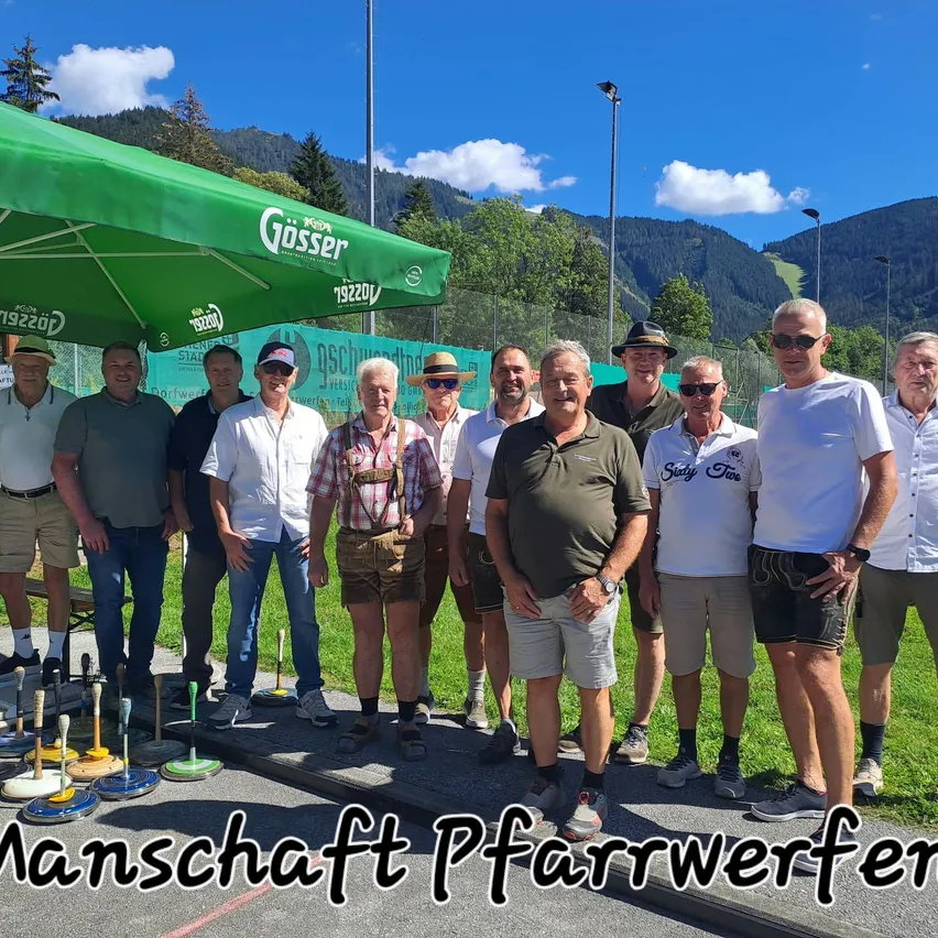 Eine Gruppe von Männern in lässiger Kleidung, einige mit Hüten, stehen unter einem grünen Schirm mit dem Wort Gosser darauf. Sie lächeln und posieren für ein Foto mit einem Grasfeld und Bergen im Hintergrund.