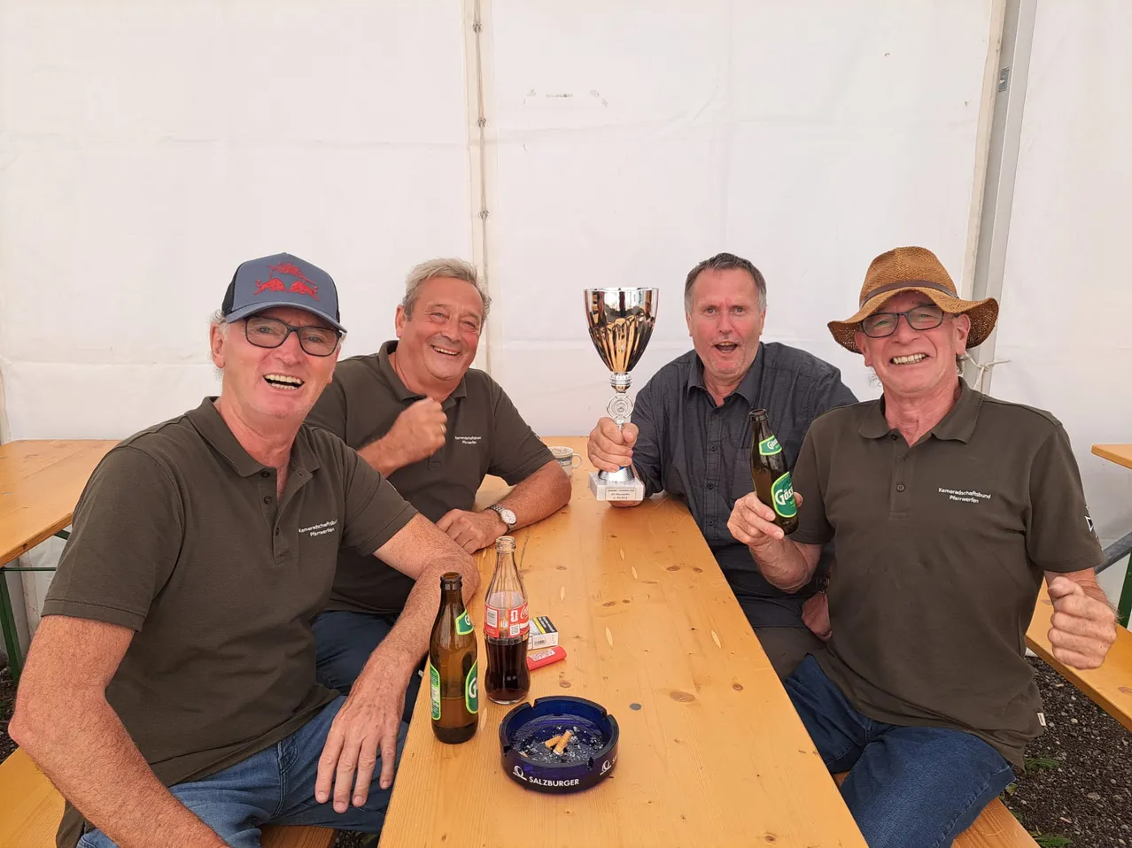 Vier Männer sitzen an einem Tisch, lächeln und posieren für ein Foto. Sie haben einen Pokal, Flaschen und eine Zigarettenasche auf dem Tisch.
