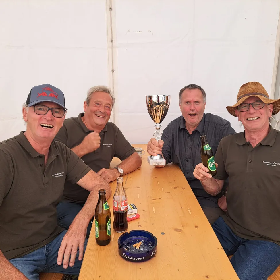 Vier Männer sitzen an einem Tisch, lächeln und posieren für ein Foto. Sie haben einen Pokal, Flaschen und eine Zigarettenasche auf dem Tisch.
