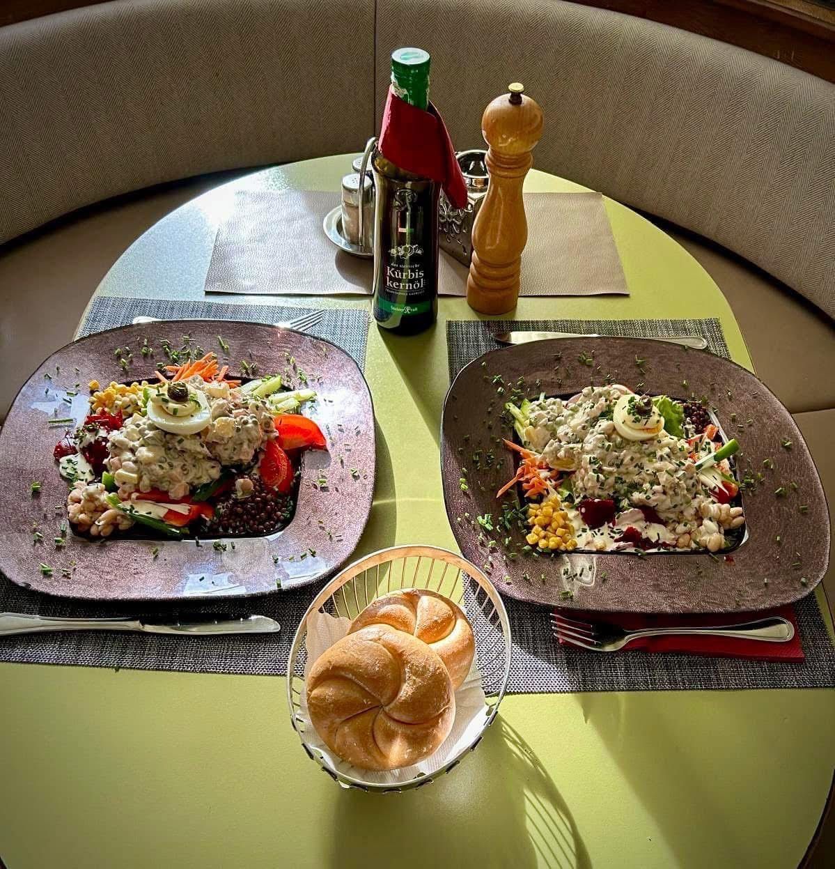 Zwei Teller mit Salat, gekochten Eiern und Brot auf einem Tisch. Eine Flasche Öl und eine Pfeffermühle stehen auf dem Tisch.