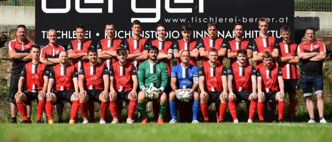 Eine Fußballmannschaft in roten und schwarzen Trikots, einige in grün, posiert für ein Gruppenfoto auf dem Rasen. Das Wort 'berger' ist auf einem schwarzen Hintergrund hinter ihnen prominent angebracht.